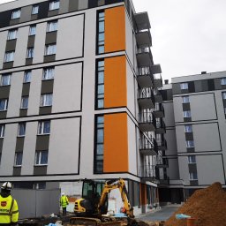 jbngroup - Nowoczesny budynek wielorodzinny w trakcie budowy, z widoczną elewacją w kolorach szarości, bieli i pomarańczu, okna zabezpieczone folią, na pierwszym planie żółta koparka i pracownicy...