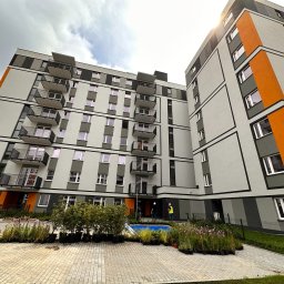 jbngroup - Nowoczesny budynek wielorodzinny z balkonami, świeżo posadzoną zielenią w donicach i dwoma pracownikami w kaskach przy wejściu.