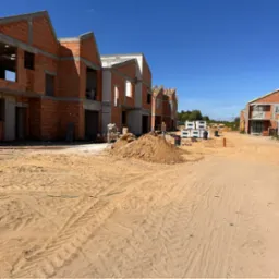 Wznoszenie szeregu domów jednorodzinnych w stanie surowym otwartym z czerwonej cegły w Warszawie, widok z piaszczystego placu budowy w słoneczny dzień.