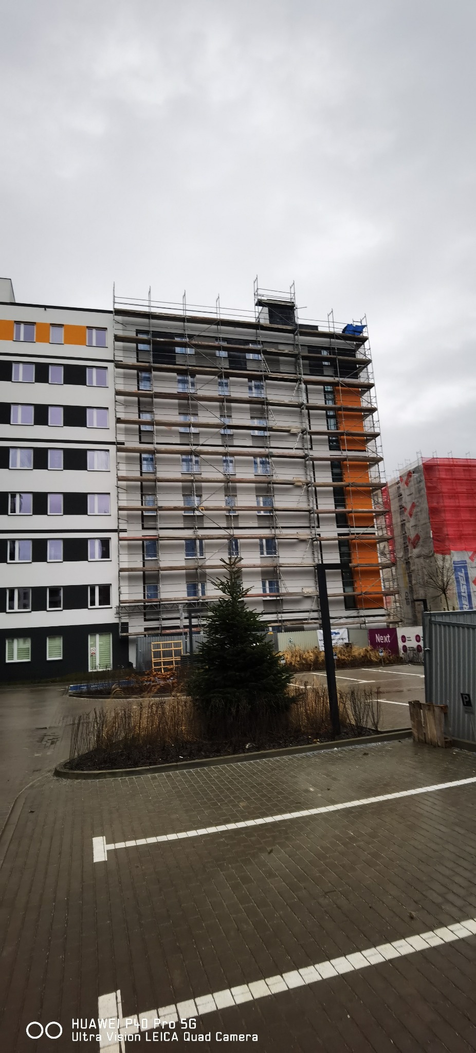 Wielopiętrowy budynek w trakcie remontu, otoczony rusztowaniami, z widocznymi fragmentami elewacji pomalowanymi na biało i pomarańczowo, na pierwszym planie małe drzewko iglaste.