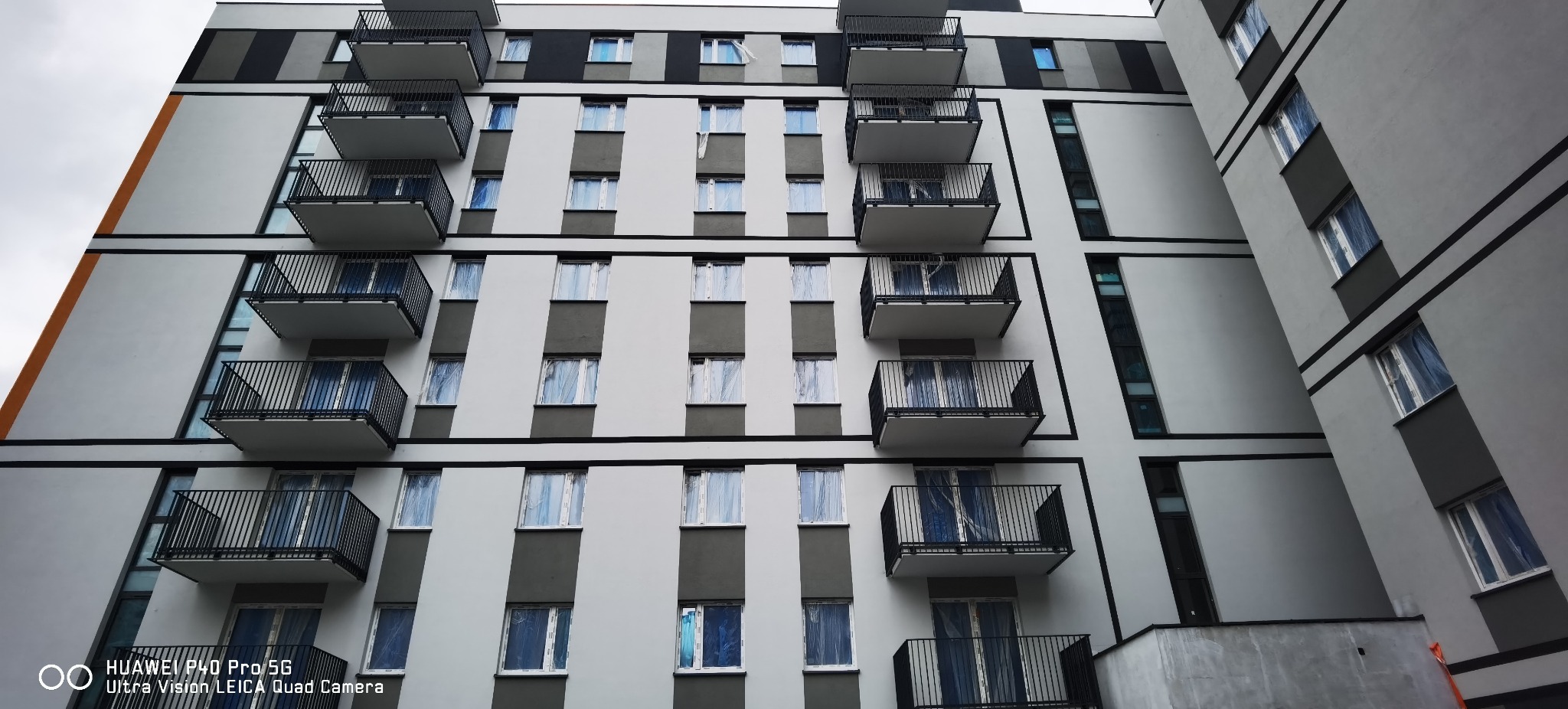 Fasada nowo wybudowanego budynku wielorodzinnego z balkonami o czarnych balustradach i oknami zabezpieczonymi folią ochronną, w kolorystyce szaro-białej.
