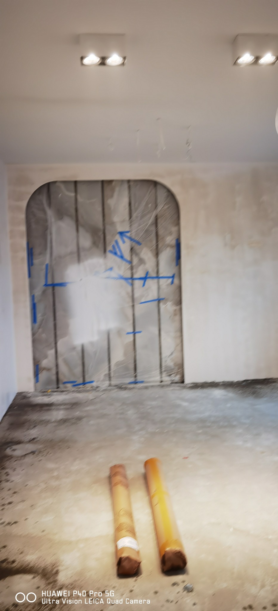 Przygotowanie do montażu płyty karton gips na ścianie z wyciętym łukiem, zabezpieczonej folią i taśmą, widoczne dwie rolki materiału na podłodze.