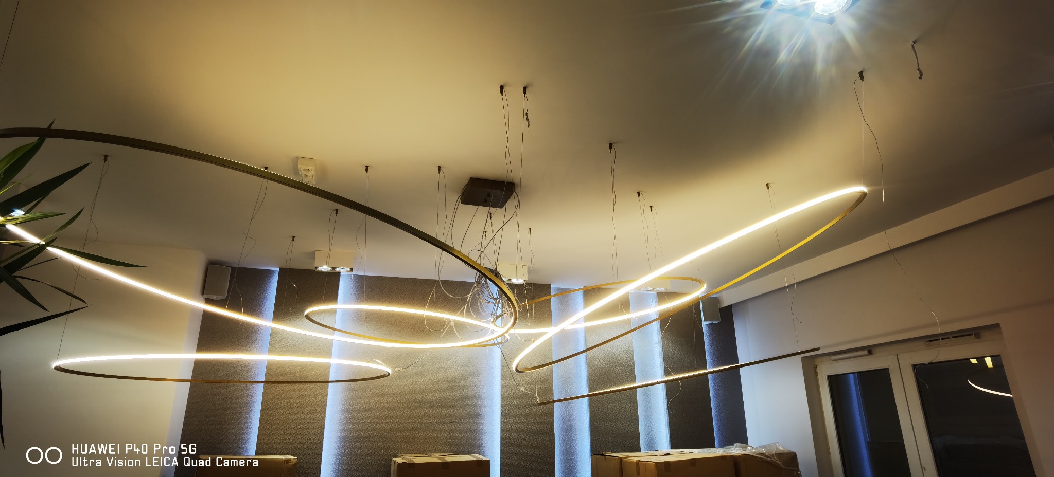 Nowoczesne oświetlenie sufitowe z podwieszanymi, okrągłymi lampami LED w złotych obudowach, widoczne kable montażowe, szary panel ścienny z pionowymi, podświetlonymi pasami, kartony na podłodze.