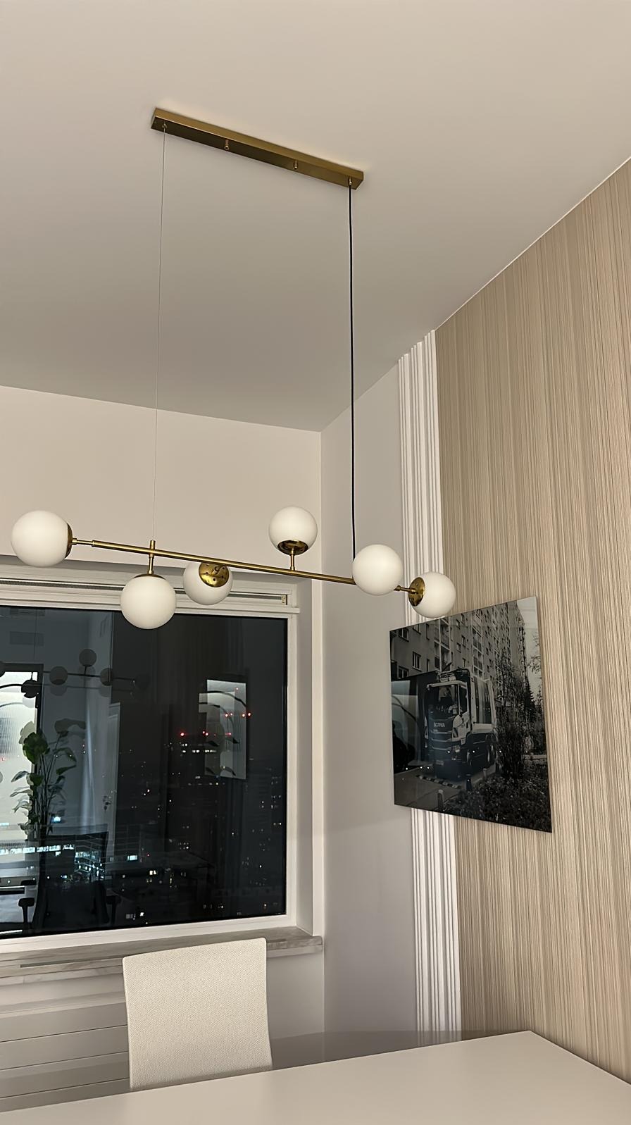 Nowoczesna lampa sufitowa z białymi kulami i złotymi detalami, zawieszona w pokoju z tapetą w pionowe paski i czarno-białym zdjęciem ciężarówki na ścianie.