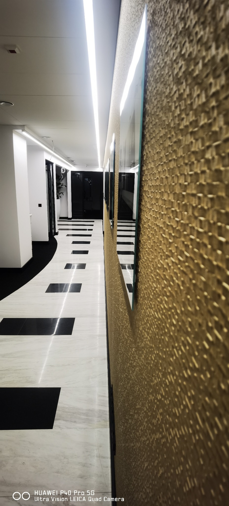 Nowoczesny korytarz biurowy z marmurową podłogą w biało-czarne kwadraty i teksturowaną, złotą ścianą z wiszącymi obrazami, oświetlony liniowymi lampami LED.