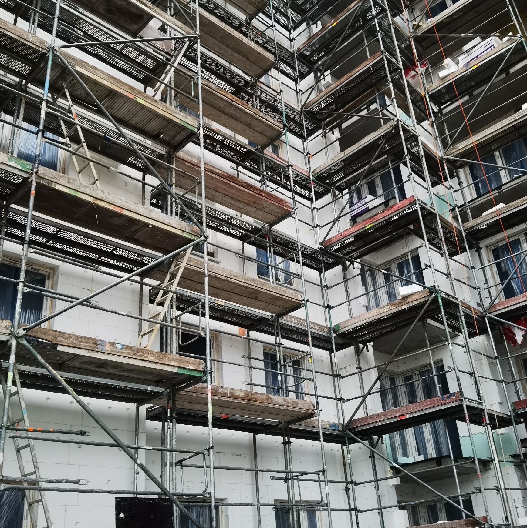 Budynek w trakcie ocieplania, obłożony białym styropianem, z rozstawionymi rusztowaniami metalowymi i drewnianymi pomostami na każdym piętrze.