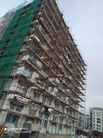 Wysoki budynek mieszkalny w trakcie prac elewacyjnych, osłonięty rusztowaniami i siatką ochronną, widoczne okna i fragmenty ocieplenia.