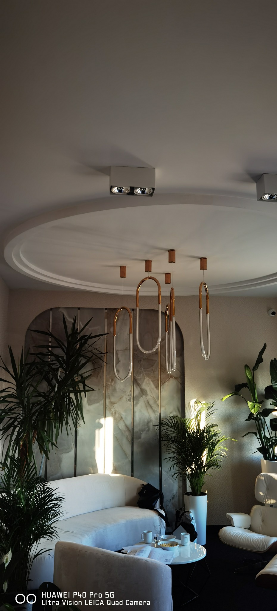 Nowoczesne wnętrze biurowe z białymi meblami, marmurową ścianą, roślinami doniczkowymi i designerską lampą wiszącą z elementami w kolorze złota.