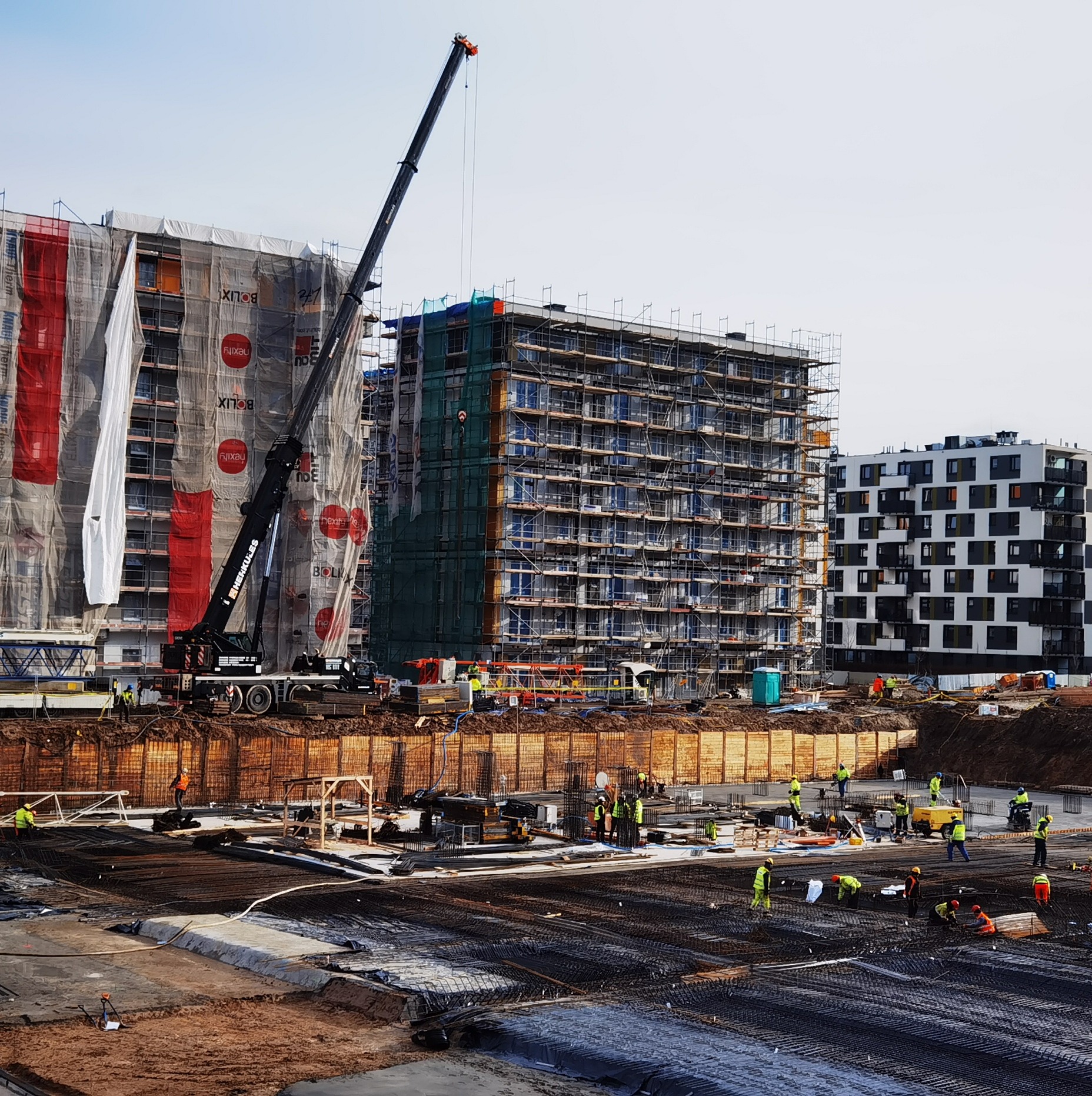 Budowa wielopiętrowego budynku mieszkalnego z rusztowaniami i dźwigiem, widoczni pracownicy w odblaskowych kamizelkach, zbrojenie fundamentów na pierwszym planie.