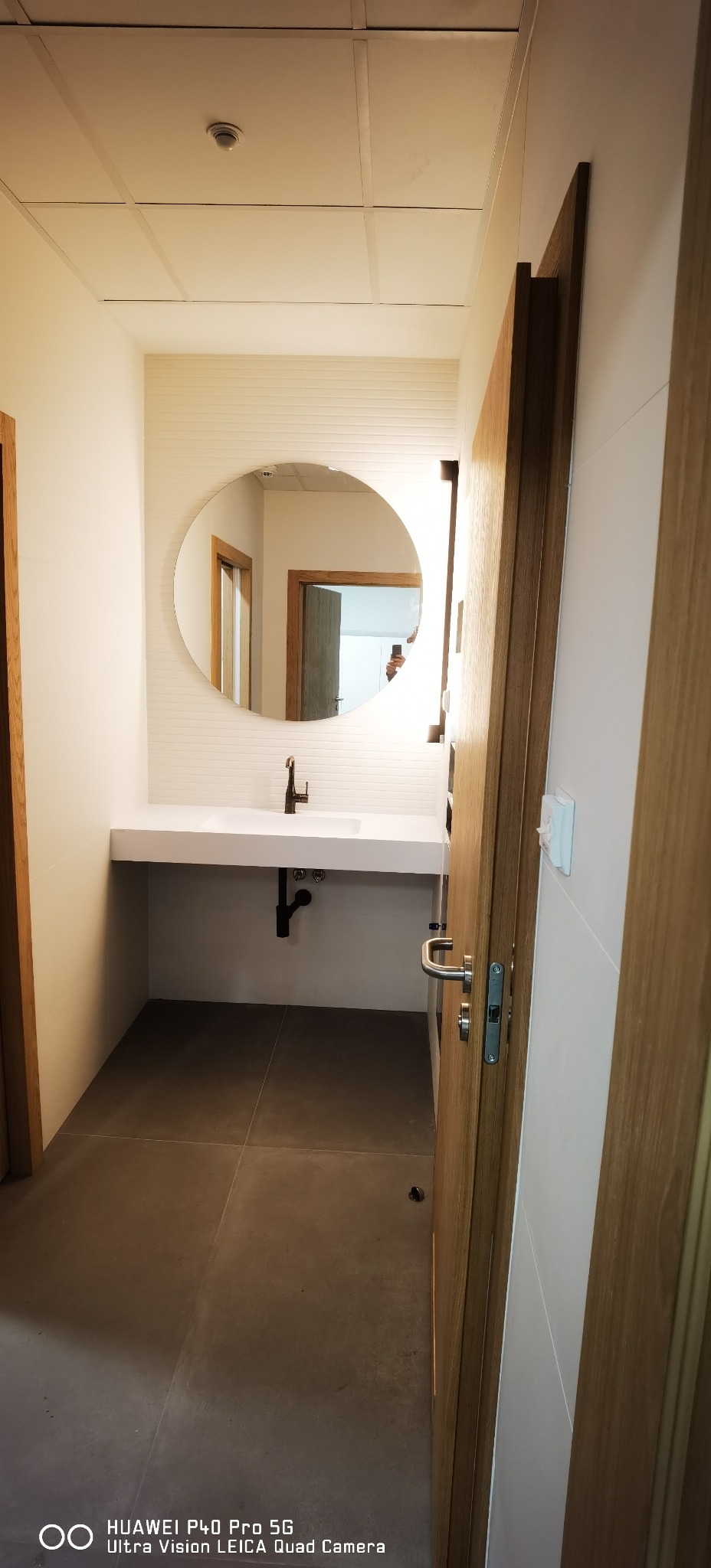 Nowoczesna łazienka z okrągłym lustrem nad umywalką, szarymi płytkami podłogowymi i drewnianymi drzwiami, widok z perspektywy wejścia.