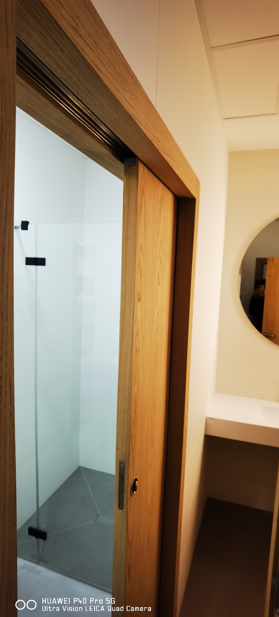 Szklana kabina prysznicowa z przesuwnymi drzwiami z jasnego drewna, widoczny fragment łazienki z okrągłym lustrem i półką