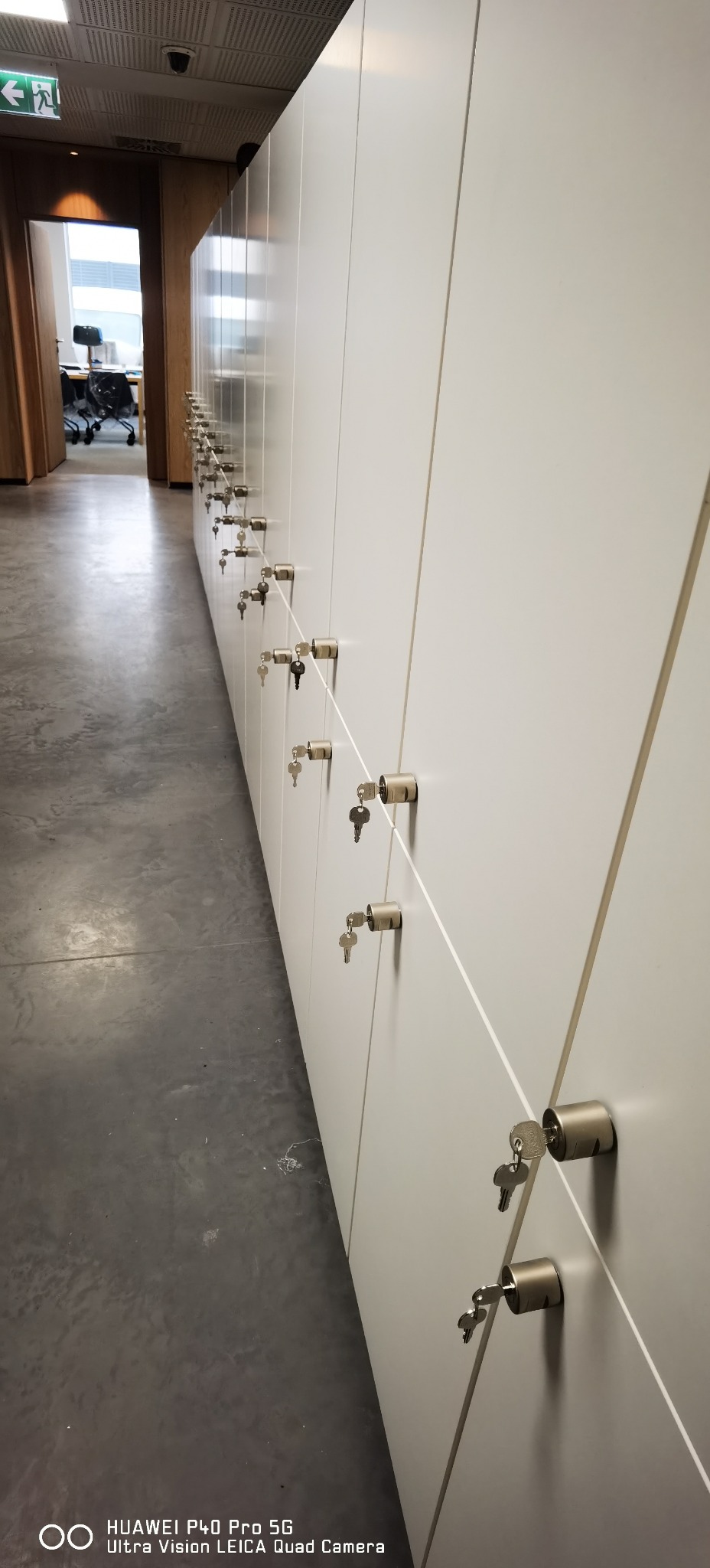 Rząd białych szafek z zamkami cylindrycznymi i kluczami w biurze, widok z perspektywy, szary beton na podłodze.