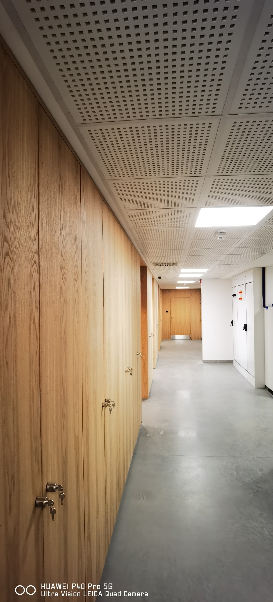Długi korytarz biurowy z rzędem drewnianych szafek zamykanych na klucz po lewej stronie i białymi szafkami po prawej, betonowa podłoga i perforowany sufit z oświetleniem.
