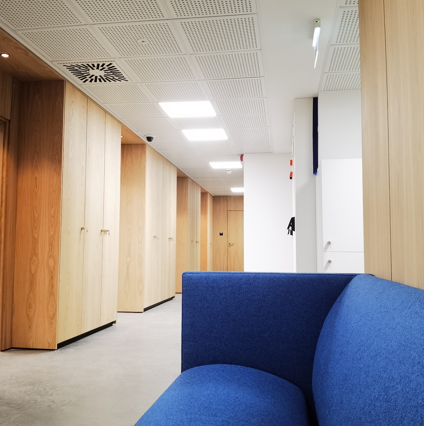 Korytarz biurowy z rzędem drewnianych szafek, widok zza niebieskiej sofy, sufit z perforowanymi panelami i oświetleniem rastrowym.