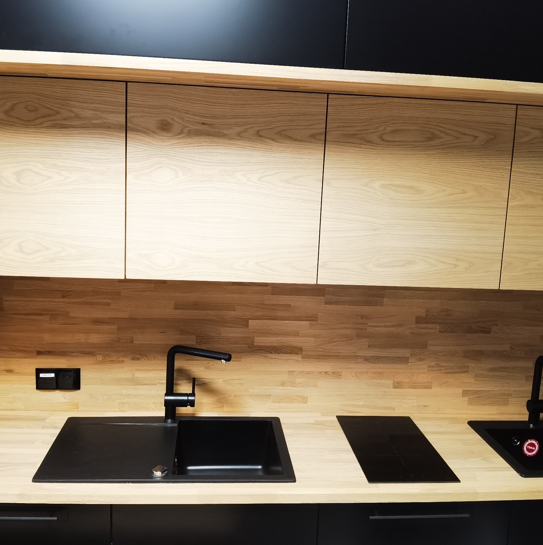 Nowoczesna kuchnia z drewnianymi szafkami górnymi i blatem z jasnego drewna, czarny zlew z baterią, płyta indukcyjna, czarne szafki dolne, drewniana mozaika na ścianie.