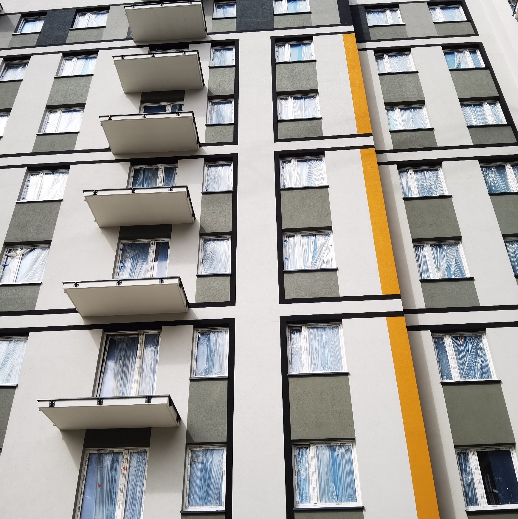 Elewacja nowo wybudowanego budynku wielorodzinnego z balkonami i oknami zabezpieczonymi folią ochronną, z elementami w kolorze szarym, białym i żółtym.