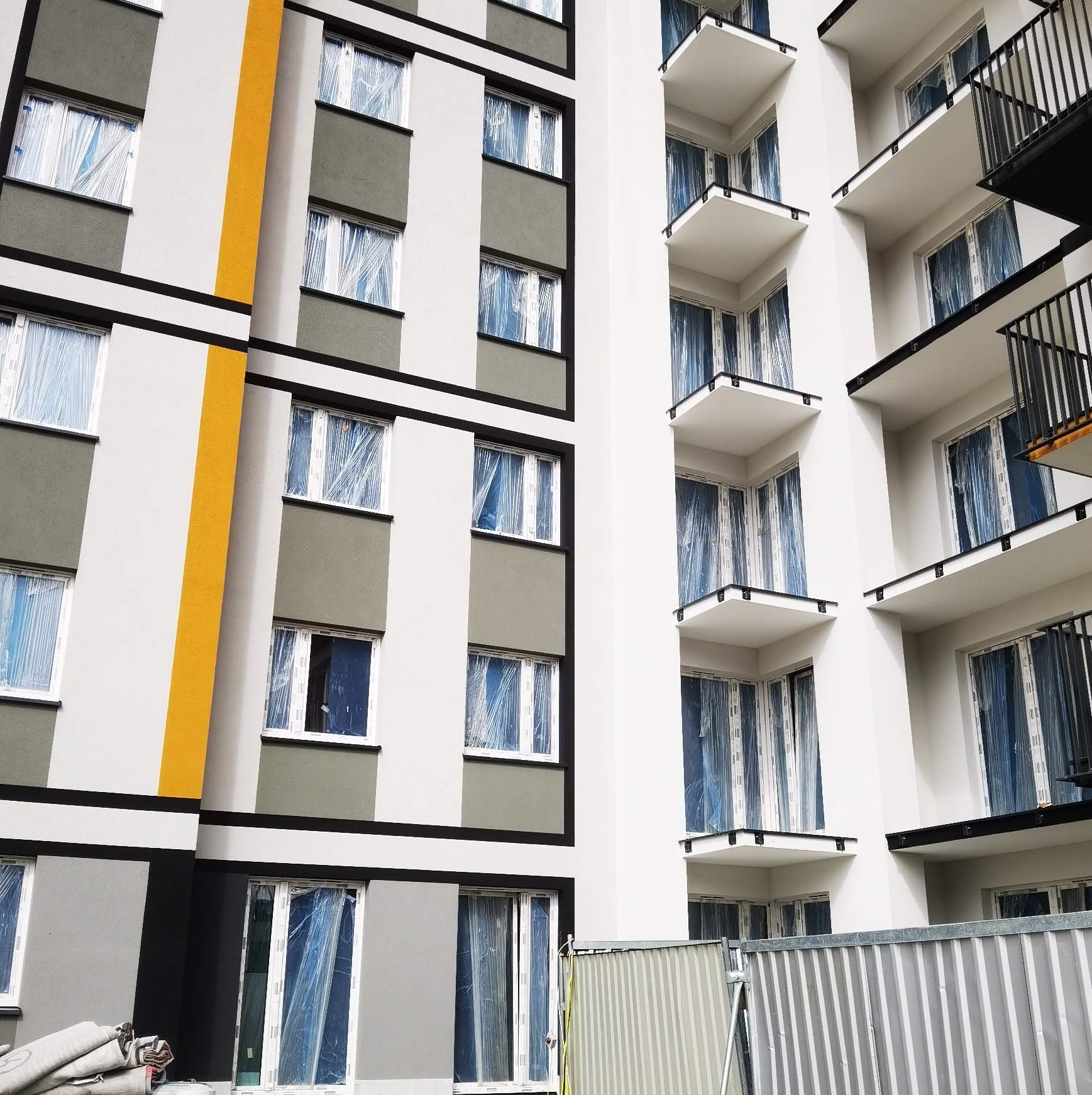 Elewacja nowego budynku wielorodzinnego z balkonami, okna zabezpieczone folią, częściowo pomalowana na szaro i biało, z żółtym akcentem, widoczne rusztowania i materiały budowlane.