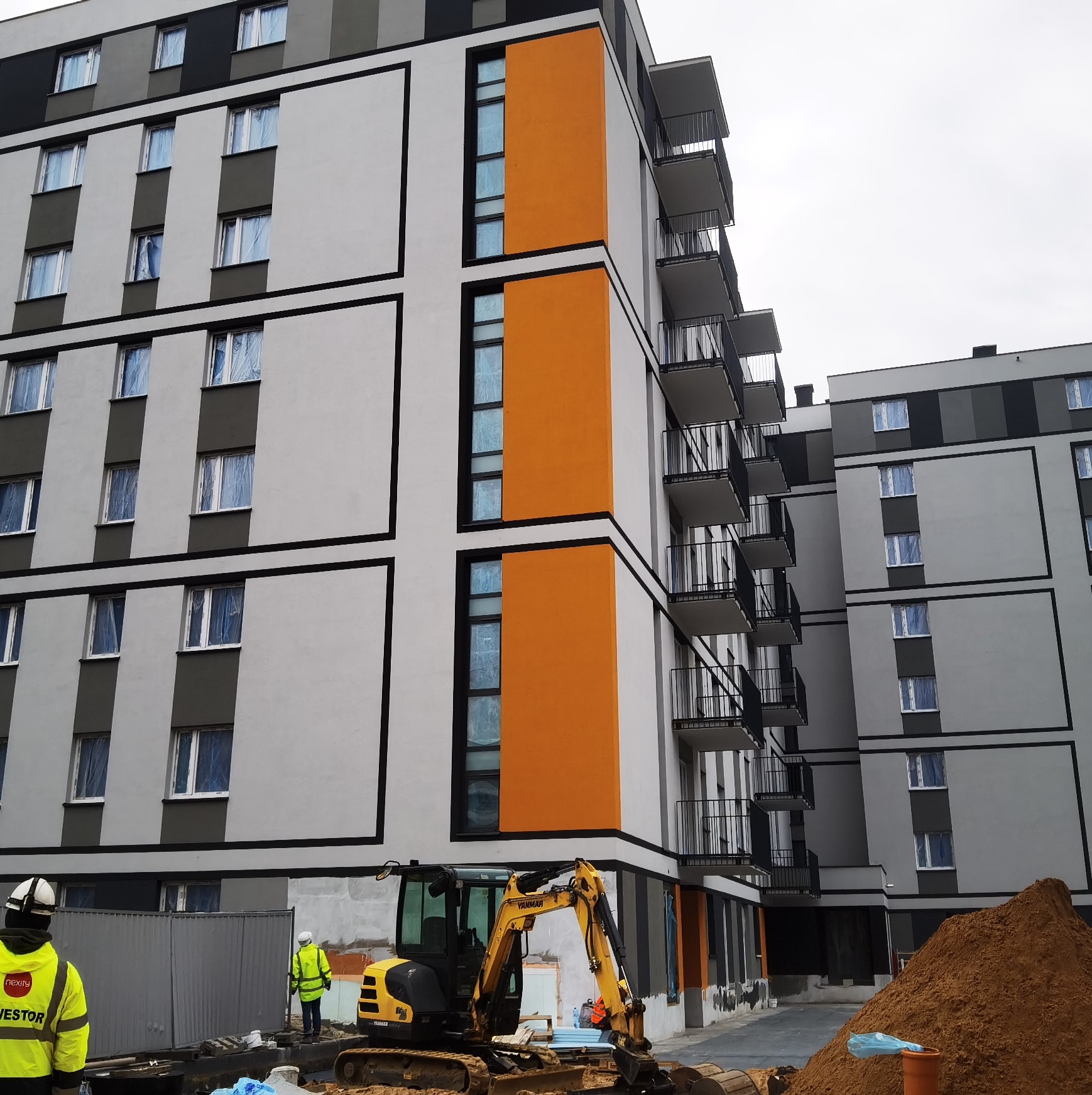 Nowoczesny budynek wielorodzinny w trakcie budowy, z widoczną elewacją w kolorach szarości, bieli i pomarańczu, okna zabezpieczone folią, na pierwszym planie żółta koparka i pracownicy...