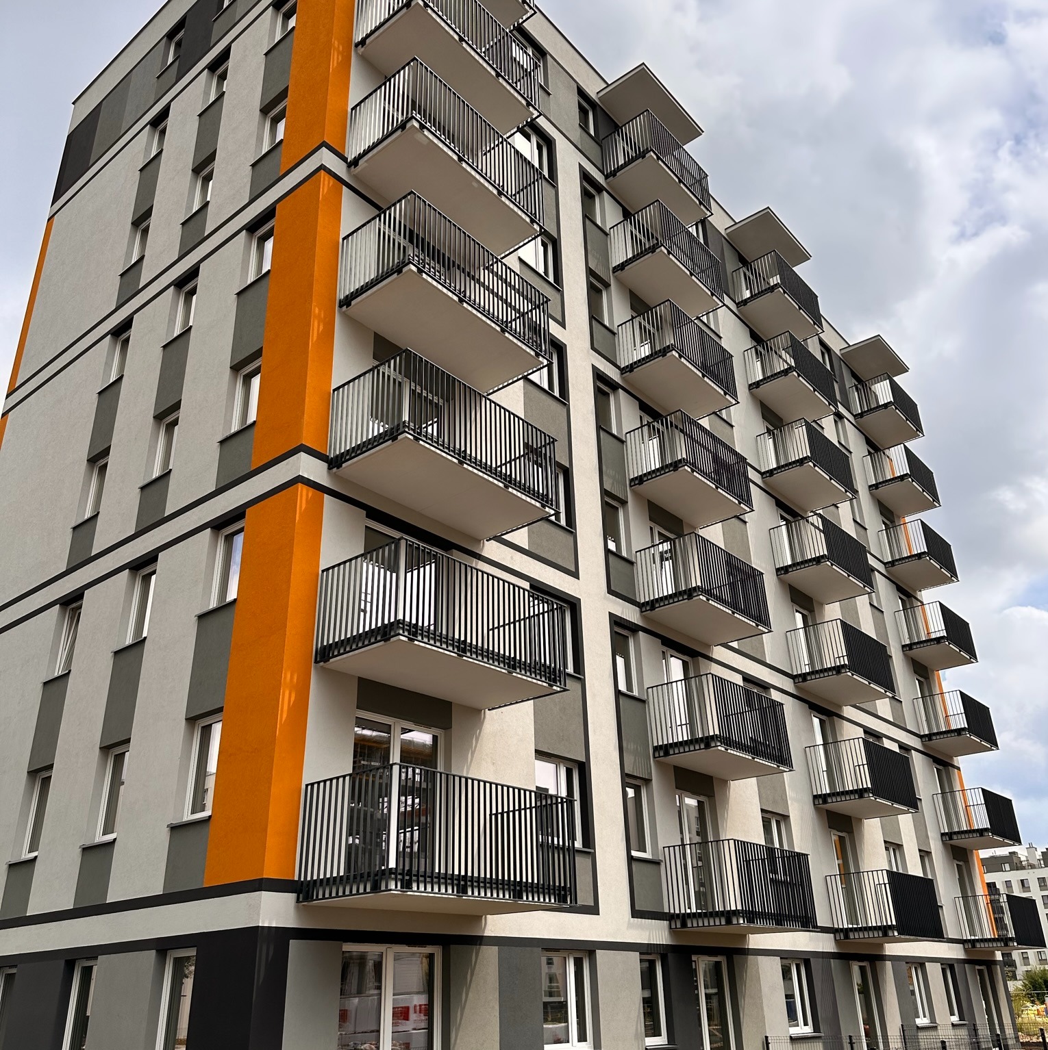 Nowoczesny budynek mieszkalny z balkonami, pomalowany w odcieniach szarości z pomarańczowym akcentem, widok z dołu na tle pochmurnego nieba.