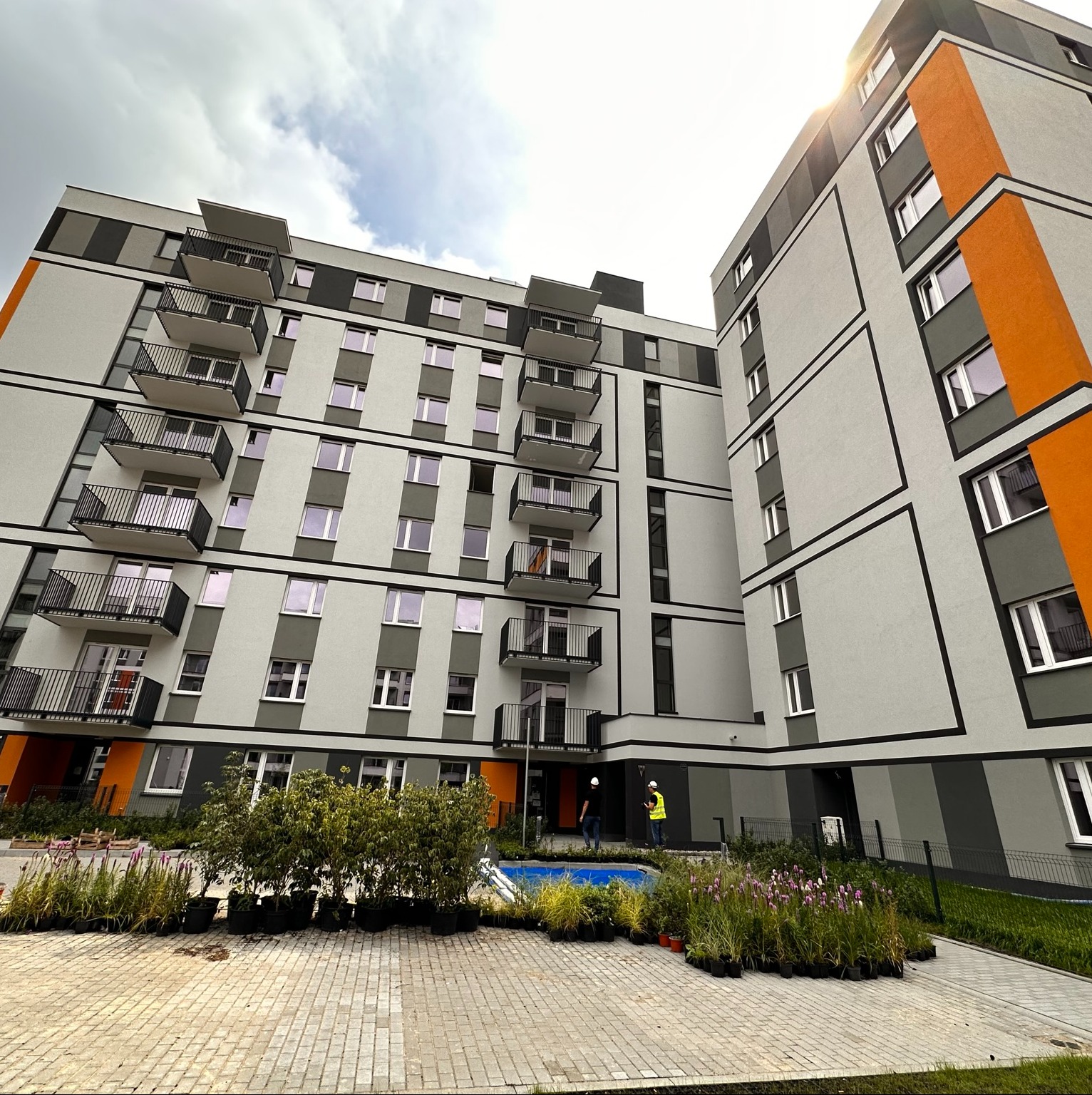 Nowoczesny budynek wielorodzinny z balkonami, świeżo posadzoną zielenią w donicach i dwoma pracownikami w kaskach przy wejściu.
