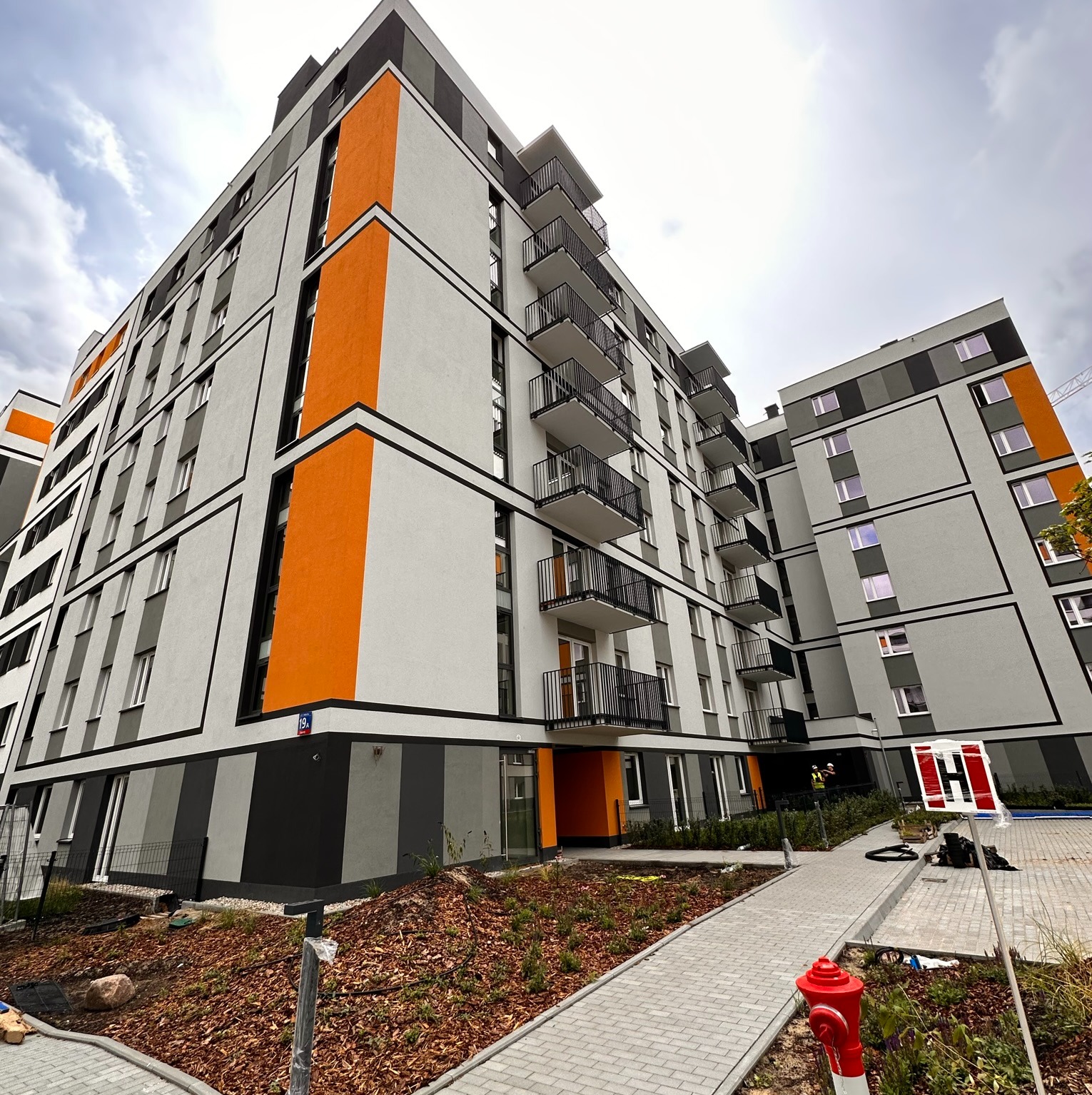 Nowoczesny budynek wielorodzinny w Warszawie z pomarańczowymi akcentami na elewacji, widok z dołu na odnowioną fasadę i balkony.