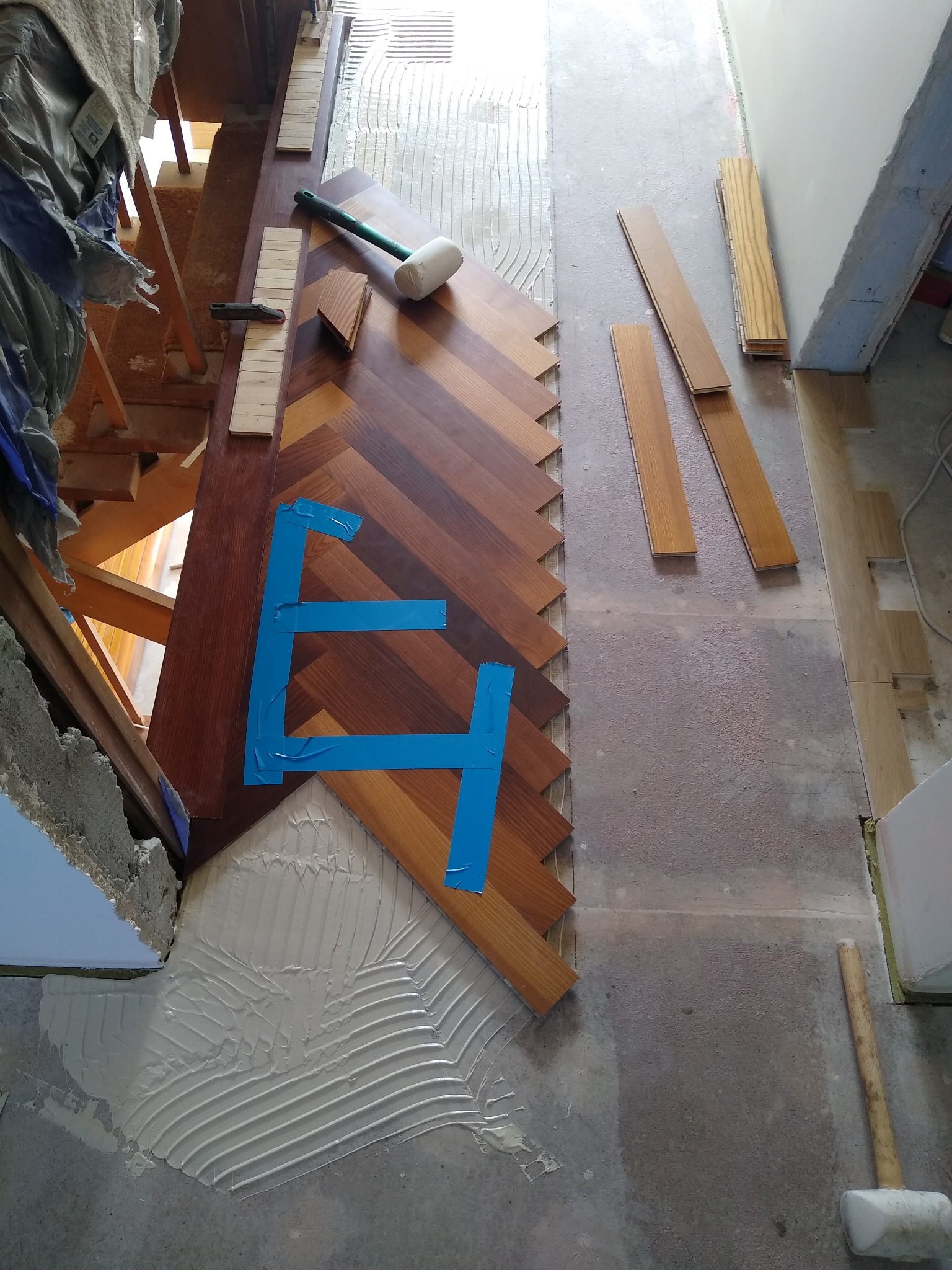 Fragment układania wzoru jodełki z drewnianych desek, zabezpieczonego niebieską taśmą, widoczne narzędzia i klej na podłożu.