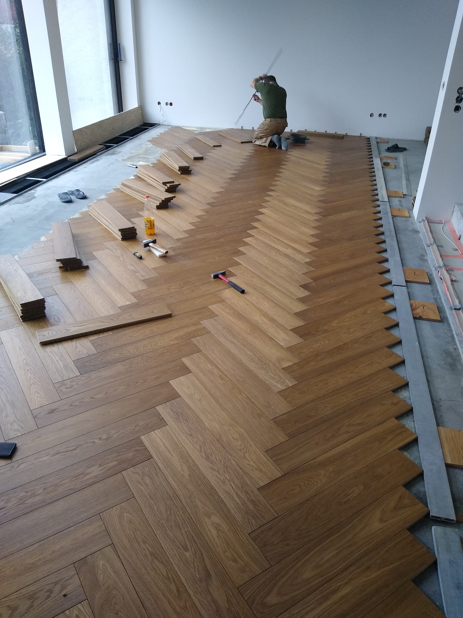 Układanie parkietu w jodełkę. Widoczny fragment ułożonej podłogi, narzędzia i pracownik w trakcie montażu. Jasne wnętrze z dużym oknem.