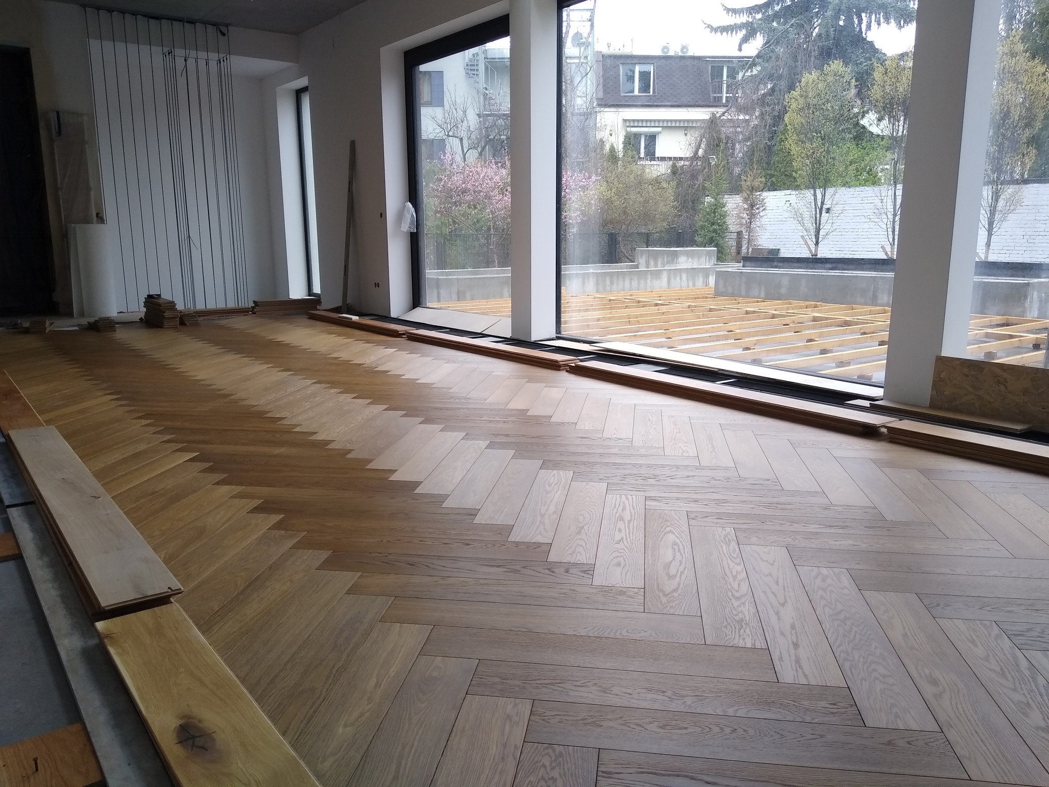 Parkiet ułożony we wzór jodełki francuskiej w jasnym, przestronnym wnętrzu z dużym oknem. Część podłogi jeszcze nieukończona, widoczne deski do ułożenia.