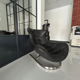 NFB INVEST SP. Z O.O. - Czarne skórzane krzesło bujane z chromowanymi nogami na błyszczącej białej podłodze w nowoczesnym biurze z czarnymi ramami okiennymi i fragmentem ściany z czerwonej cegły.