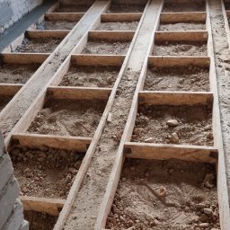 NFB INVEST SP. Z O.O. - Konstrukcja drewniana pod podłogę wypełniona piaskiem i gruzem, widoczne ślady butów, surowy mur z cegły, okno w tle.