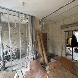 NFB INVEST SP. Z O.O. - Wnętrze w trakcie remontu: konstrukcja metalowa pod płytę karton gips na tle starej ceglanej ściany, widoczne materiały budowlane, drabina i lampa wisząca.