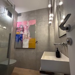 NFB INVEST SP. Z O.O. - Nowoczesna łazienka z szarą płytką, wiszącą toaletą za szklaną kabiną, białą umywalką nablatową, abstrakcyjnym obrazem na ścianie i designerską lampą wiszącą z kulistymi kloszami.