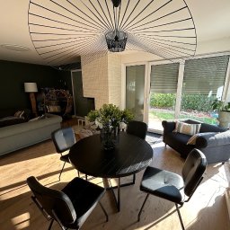 NFB INVEST SP. Z O.O. - Nowoczesne wnętrze domu z okrągłym, czarnym stołem jadalnym, czarnymi krzesłami i designerską lampą sufitową w kształcie wachlarza, oświetlone promieniami słońca padającymi przez okno.