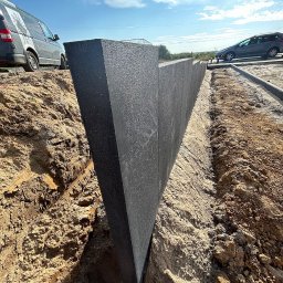 NFB INVEST SP. Z O.O. - Czarne, trójkątne panele z tworzywa sztucznego wkopane w ziemię, tworzące ogrodzenie lub zabezpieczenie terenu budowy, widoczne samochody w tle.