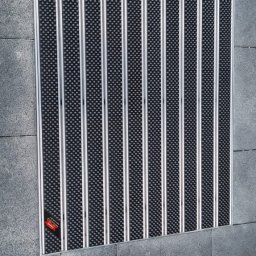 NFB INVEST SP. Z O.O. - Wpuszczona wycieraczka systemowa z aluminiowymi profilami i czarnym wkładem czyszczącym, zamontowana w nawierzchni z kostki brukowej w Szczecinie.