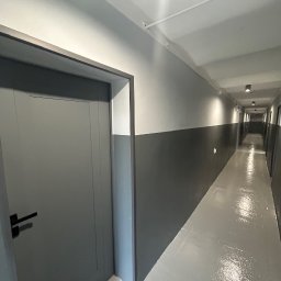 NFB INVEST SP. Z O.O. - Korytarz w budynku po remoncie, ściany pomalowane na szaro z dwoma odcieniami, widoczne drzwi z czarną klamką, podłoga szara, oświetlenie sufitowe.