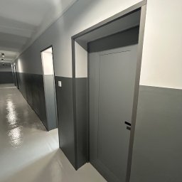 NFB INVEST SP. Z O.O. - Szary korytarz biurowy po remoncie z nowymi drzwiami i pomalowanymi ścianami na biało i szaro, widoczne lampy sufitowe.