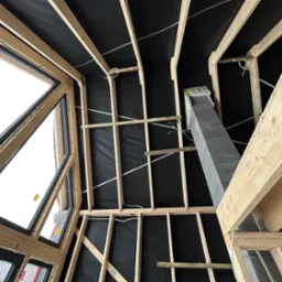 Wnętrze poddasza w trakcie ocieplania: widoczna konstrukcja drewniana, czarna membrana, komin z bloczków betonowych i okna o nietypowym kształcie.