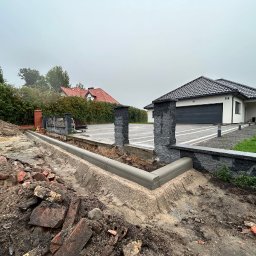 NFB INVEST SP. Z O.O. - Układanie obrzeży chodnikowych z szarego betonu wzdłuż posesji z brukowanym podjazdem i kamiennymi słupkami ogrodzeniowymi, widoczne gruz i prace ziemne.