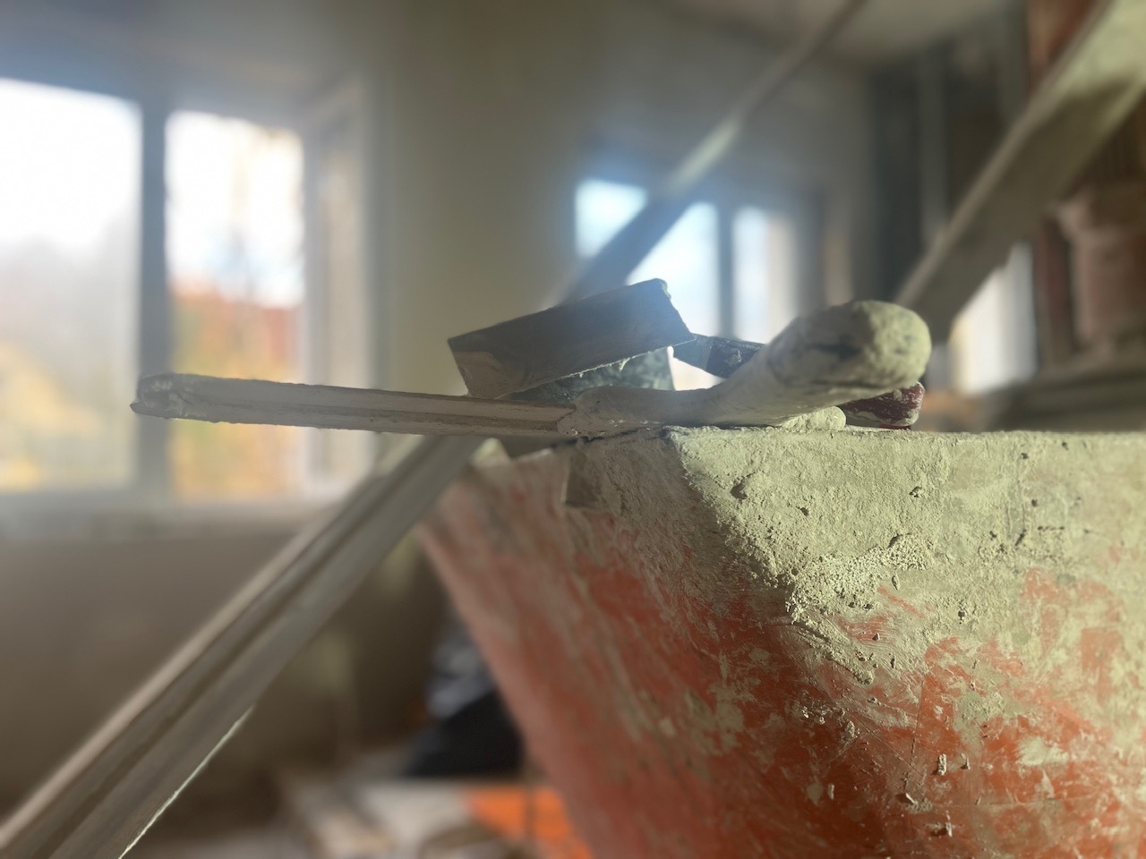 Używane narzędzia szpachlarskie leżące na krawędzi pomarańczowego wiadra z resztkami gładzi, widok z dołu, okno w tle.