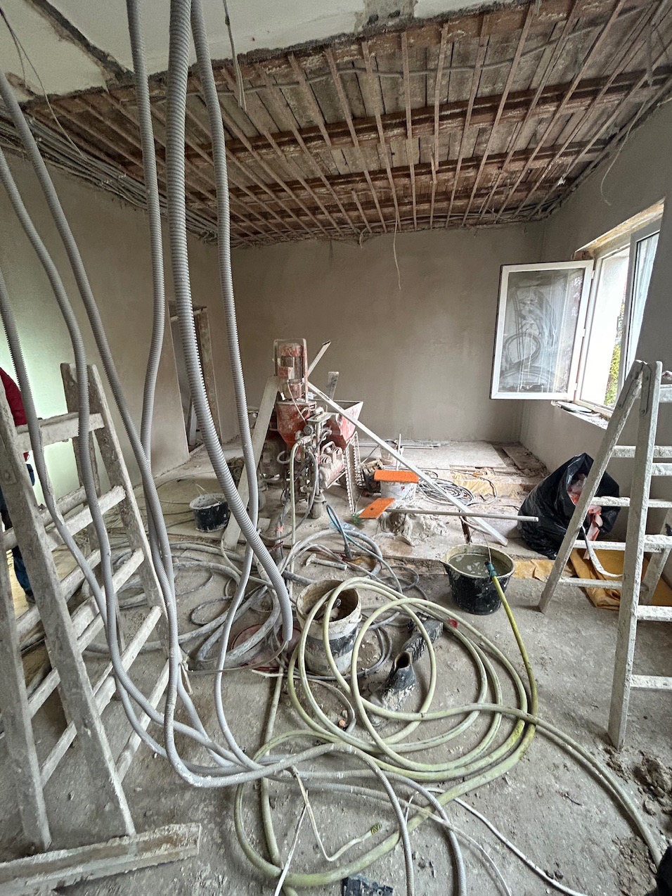 Wnętrze remontowanego pokoju z widoczną maszyną do szpachlowania, przewodami, drabinami i odsłoniętą konstrukcją sufitu.