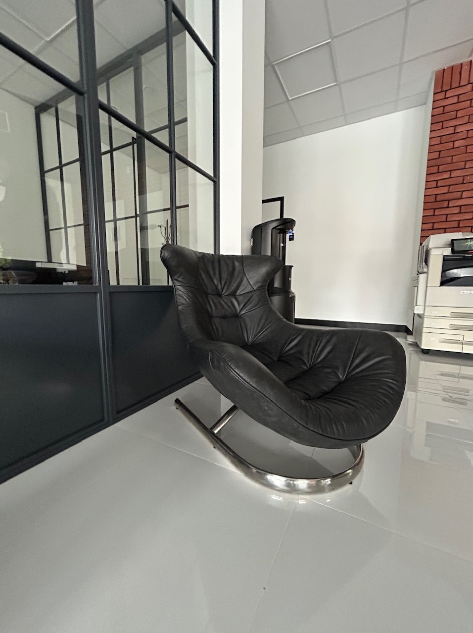 Czarne skórzane krzesło bujane z chromowanymi nogami na błyszczącej białej podłodze w nowoczesnym biurze z czarnymi ramami okiennymi i fragmentem ściany z czerwonej cegły.