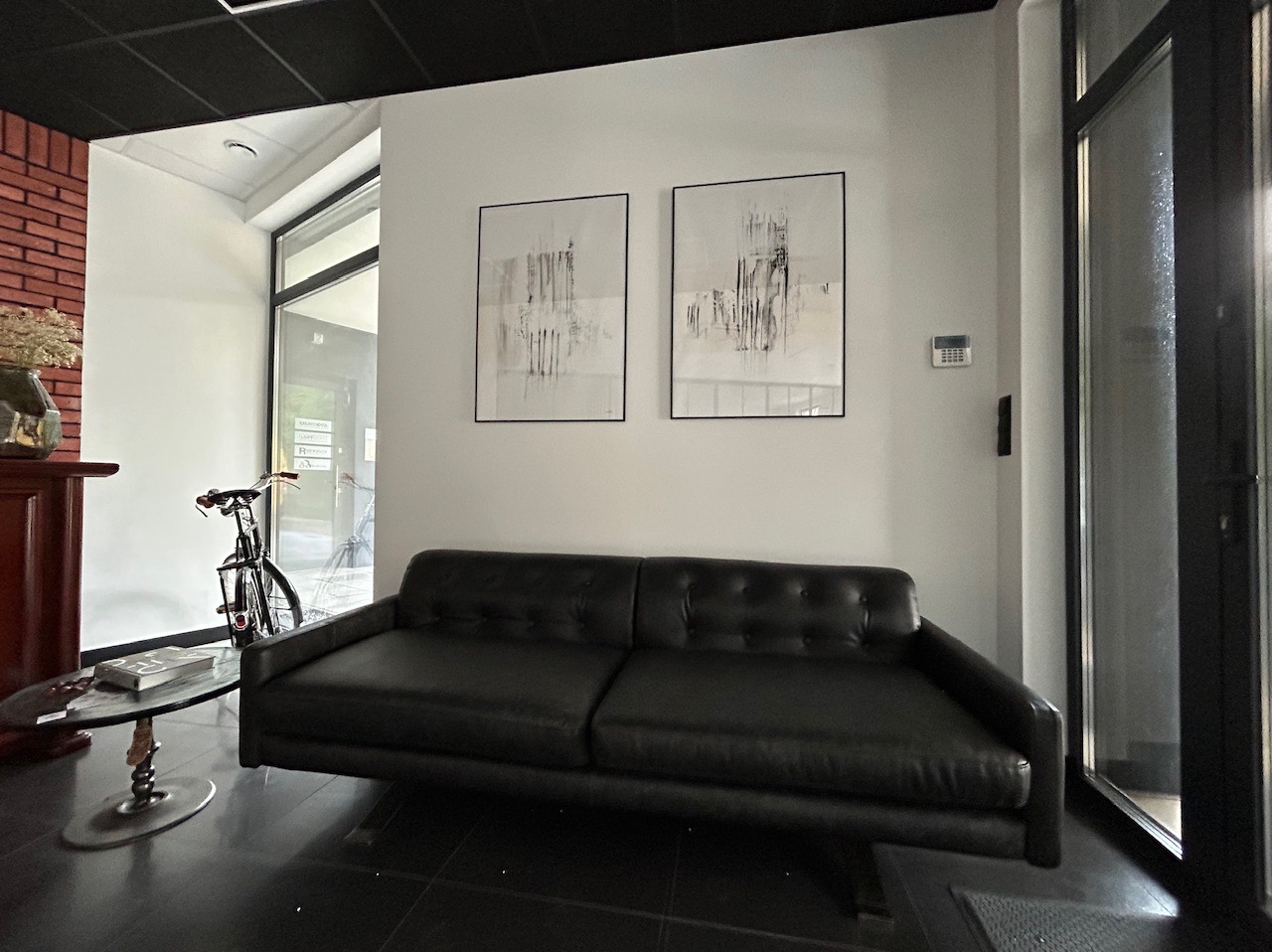 Czarne skórzane dwuosobowe siedzisko pod dwoma obrazami w czarnych ramkach w jasnym pomieszczeniu z rowerem widocznym w tle za szklanymi drzwiami.