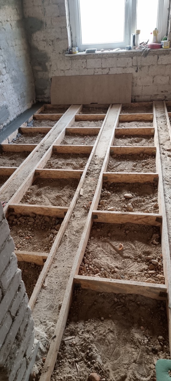 Konstrukcja drewniana pod podłogę wypełniona piaskiem i gruzem, widoczne ślady butów, surowy mur z cegły, okno w tle.