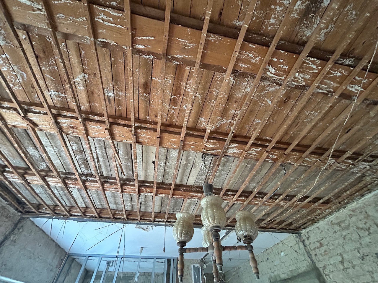 Drewniana konstrukcja sufitu z widocznymi belkami i śladami po usuniętej płycie, w tle fragment ściany z cegły i wisząca lampa.