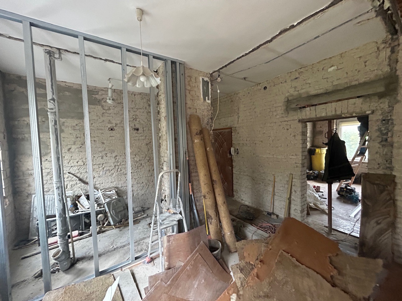 Wnętrze w trakcie remontu: konstrukcja metalowa pod płytę karton gips na tle starej ceglanej ściany, widoczne materiały budowlane, drabina i lampa wisząca.