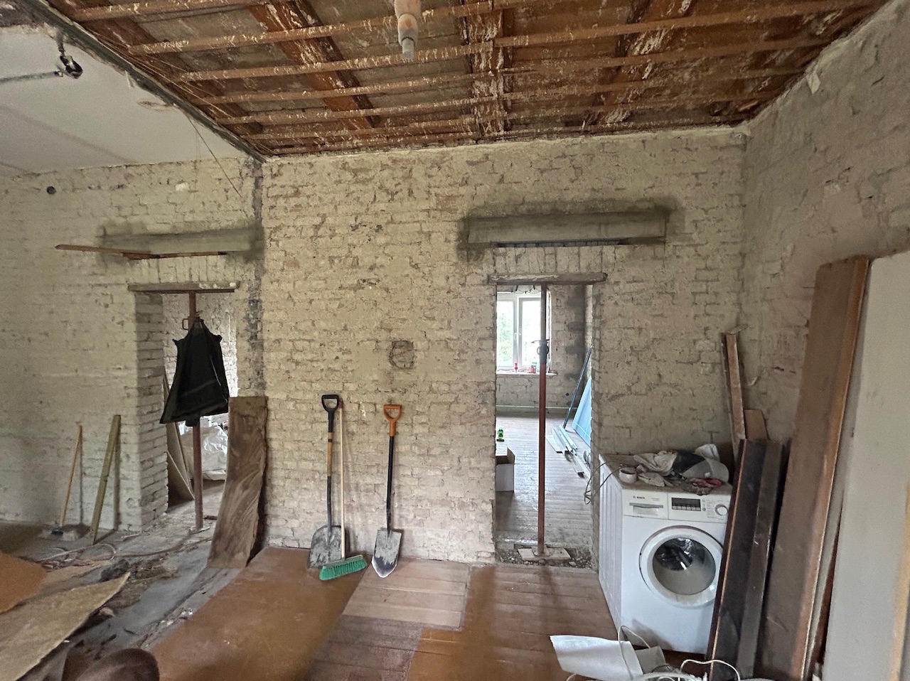 Wnętrze w trakcie remontu: odsłonięta cegła, drewniany strop, narzędzia (szpadle i miotła), pralka w pomieszczeniu, widoczne ślady prac budowlanych.