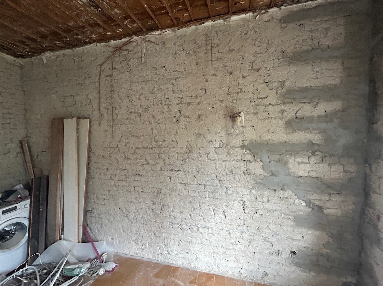 Wnętrze w trakcie prac, ściana z cegły pokryta częściowo szpachlą, widoczne instalacje na suficie, podłoga z paneli, pralka i materiały budowlane przy ścianie.