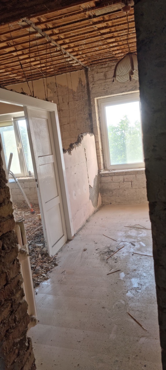 Wnętrze pokoju w trakcie demontażu, widoczne odsłonięte cegły, resztki tynku na ścianach, drewniany strop z wystającymi przewodami, stare okno i drzwi, gruz na podłodze.