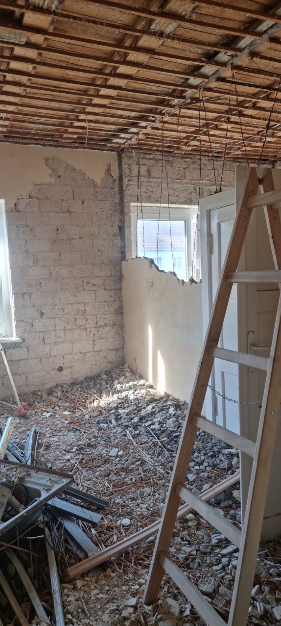 Wnętrze w trakcie demontażu: odsłonięta cegła, resztki tynku na ścianach, drewniany strop z wiszącymi przewodami, gruz na podłodze, drabina oparta o framugę drzwi.